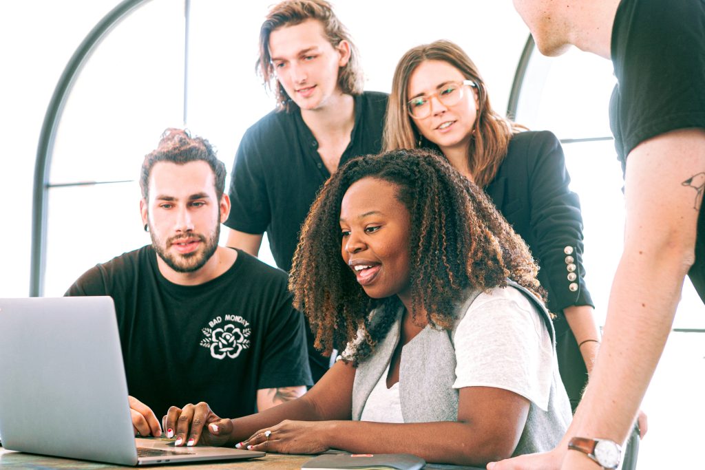 Four diverse colleagues collaborate eagerly around a laptop in a modern office, engaged in a discussion, showing teamwork and focus.