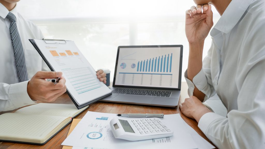 Two professionals in a meeting discussing charts and data on a laptop and paper documents, with calculators and other stationery items on the table.