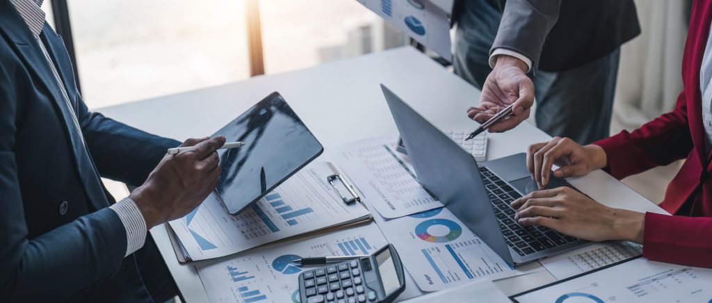 Business professionals sitting at a table, reviewing financial reports and data on laptops and digital tablets during a meeting - the desk scattered with paper documents and charts.