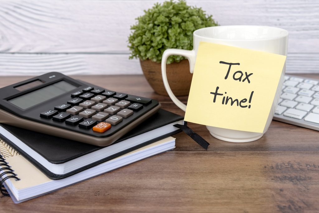 A desk setup featuring a calculator, a coffee mug with a sticky note saying "tax time!", a notebook, and a partial view of a keyboard, all on a wooden table with a green plant in the background.