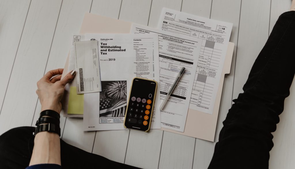 A person sits on a wooden floor reviewing tax documents and calculations on paper, with a smartphone displaying a calculator app and a pen beside them.