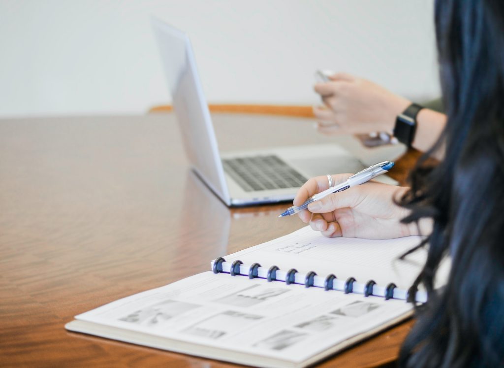 Two people sitting at a table, one writing in a notebook and the other checking their phone while sitting at a laptop.