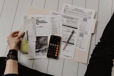 A person sits on a wooden floor reviewing tax documents and calculations on paper, with a smartphone displaying a calculator app and a pen beside them.
